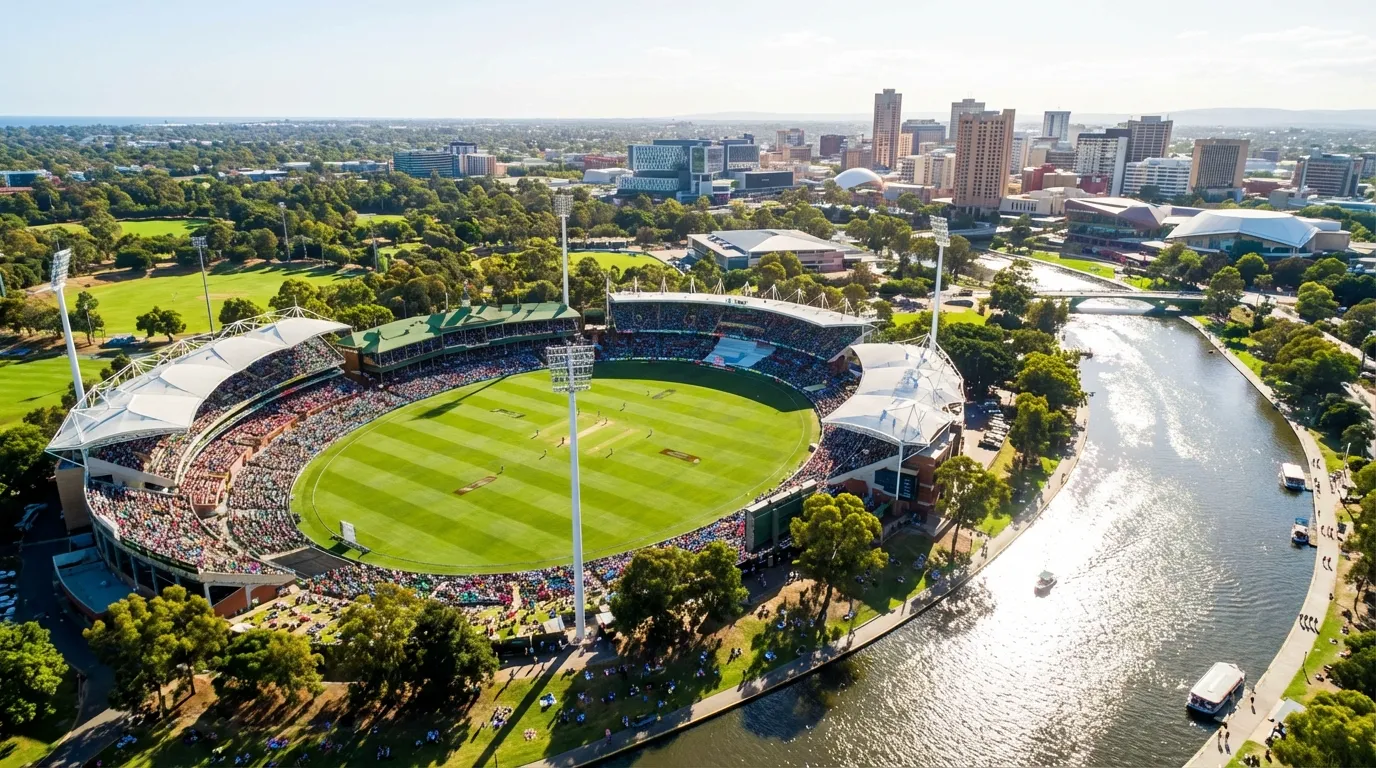 Image: A vibrant, aerial drone shot of Adelaide Oval on a sunny day, showcasing its lush green pitch, grandstands filled with a diverse crowd, and the seamless integration with the surrounding green parklands and the glistening River Torrens. The Adelaide city skyline is visible in the background, reinforcing its urban connection.