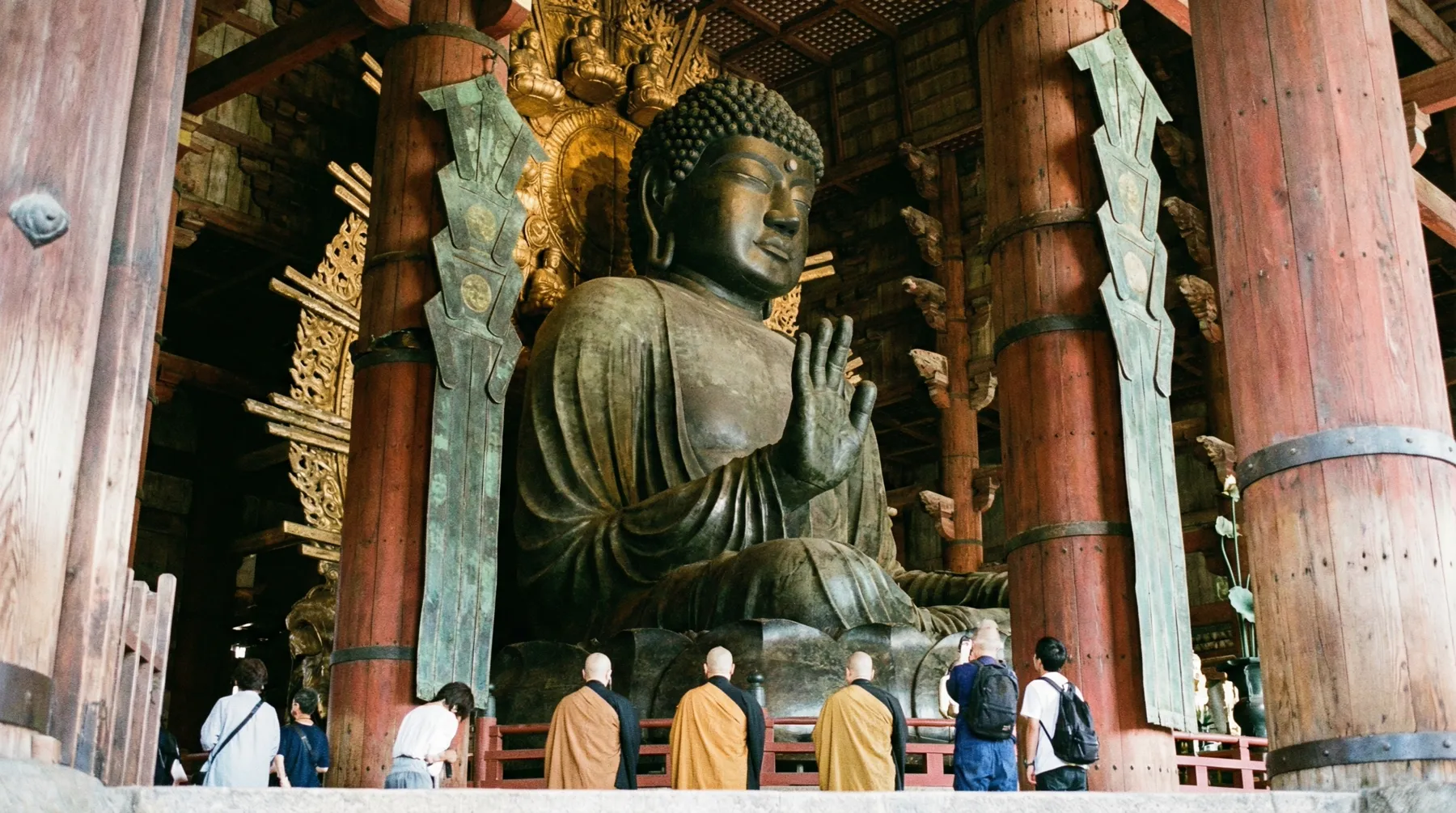 Image: A large bronze statue of the Great Buddha (Daibutsu) inside the Todai-ji Temple in Nara, Japan. The statue is lit by natural light filtering through the temple entrance, with a few respectful visitors in the foreground.
