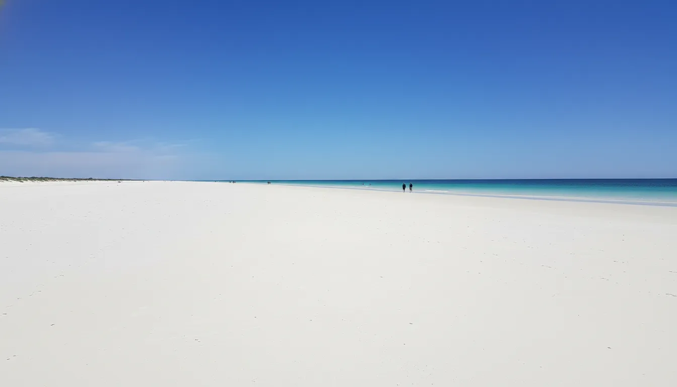 Image: A wide panoramic shot of Cable Beach at midday, showing pristine white sand stretching into the distance, turquoise Indian Ocean waters, and a clear blue sky. A few people are walking far in the distance, emphasizing the vastness.