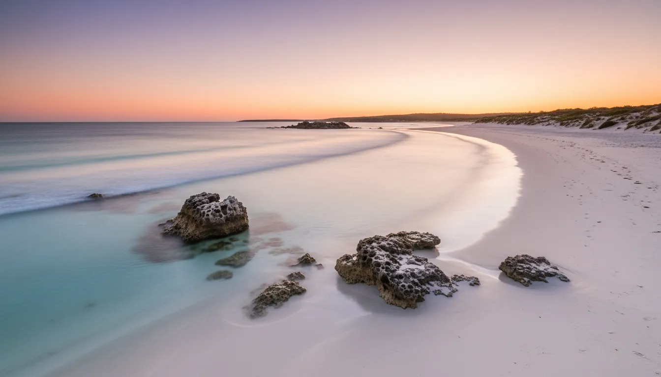 Image: A panoramic shot of a pristine, secluded beach on Rottnest Island at sunset. The sky is a gradient of soft orange, pink, and purple, reflecting on the calm, clear water. White sand stretches into the distance, with a few rugged coastal rocks. No people are visible, emphasizing the untouched natural beauty and tranquility.